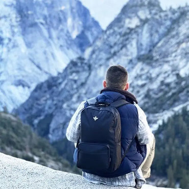 A person with an Erozul Legend Smell Proof Back Pack With Combination Lock by Erozul and a blue vest sits on a rock, looking out at snow-covered mountains and the distant valley.
