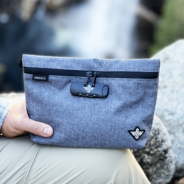 A person holds an Erozul Orion Smell Proof Bag With Combination Lock 11—a grey, water-resistant pouch with black zipper and clip—while sitting outdoors near rocks and water, perfect for outdoor adventures.