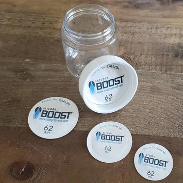 An empty clear dispensary jar and four Integra Boost Humidity Cap Pack 45mm 62% by Integra Boost are shown on a wooden surface, ready to keep your contents fresh.