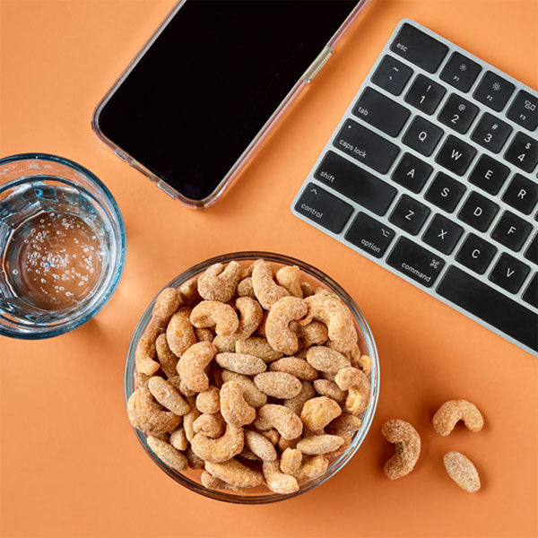 A 2oz bag of Planters Nut Duos Buffalo Cashews & Ranch Almonds, a glass of water, a smartphone, and part of a keyboard arranged on an orange surface.