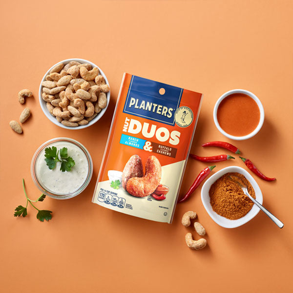 A 2oz bag from the 30 Pack - Planters Nut Duos Buffalo Cashews & Ranch Almonds sits on an orange surface, surrounded by creamy cashews, ranch dip, chili powder, red chili peppers, and a bowl of orange sauce.