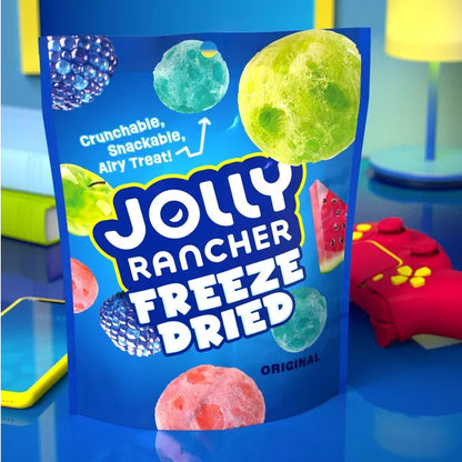 A 2oz blue bag from the 6 Pack - Jolly Rancher Freeze Dried Original Fruit Flavored Candy sits on a table by a yellow phone and red game controller, showing colorful images of these airy, crunchy fruity treats.