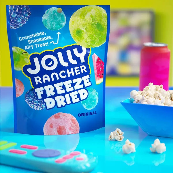A 6 Pack of Jolly Rancher Freeze Dried Original Fruit Flavored Candy 2oz Bags sits on a blue table with popcorn, a pink drink can, and a colorful remote—perfect crunchy, fruity snacks from Jolly Rancher for your next movie night.