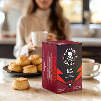 A 6 Pack of Death Wish Coffee Dark Roast Single-Serve Pods sits on a counter in front of scones, while a woman enjoys a cup of premium Death Wish coffee in the background.