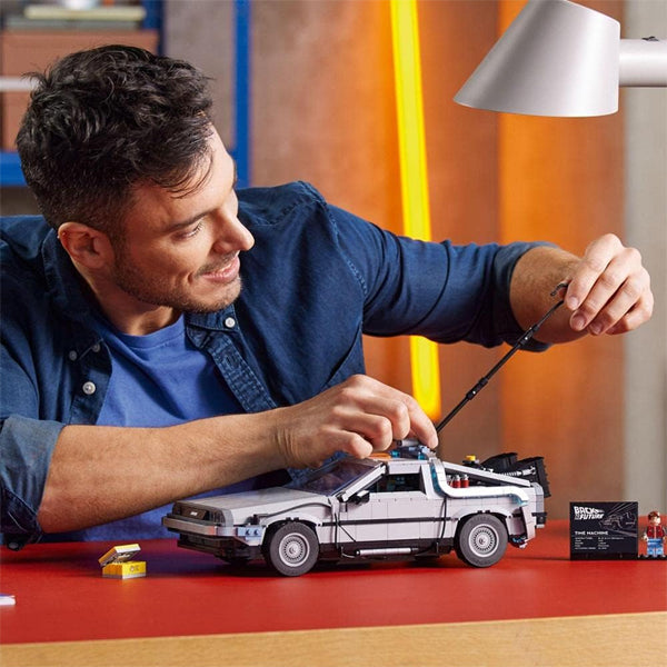 A man assembles the LEGO Back to the Future DeLorean DMC-12 10300 at a table, with an info plaque beside it, enjoying this LEGO model kit for adults and adding to his Back to the Future collection.