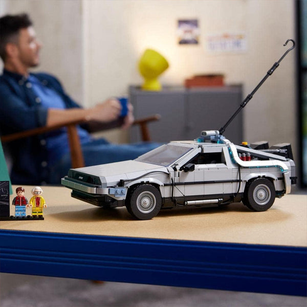 A LEGO Back to the Future DeLorean DMC-12 10300 model by LEGO is displayed on a table with two minifigures, part of Back to the Future memorabilia, while a blurred person sits in the background.